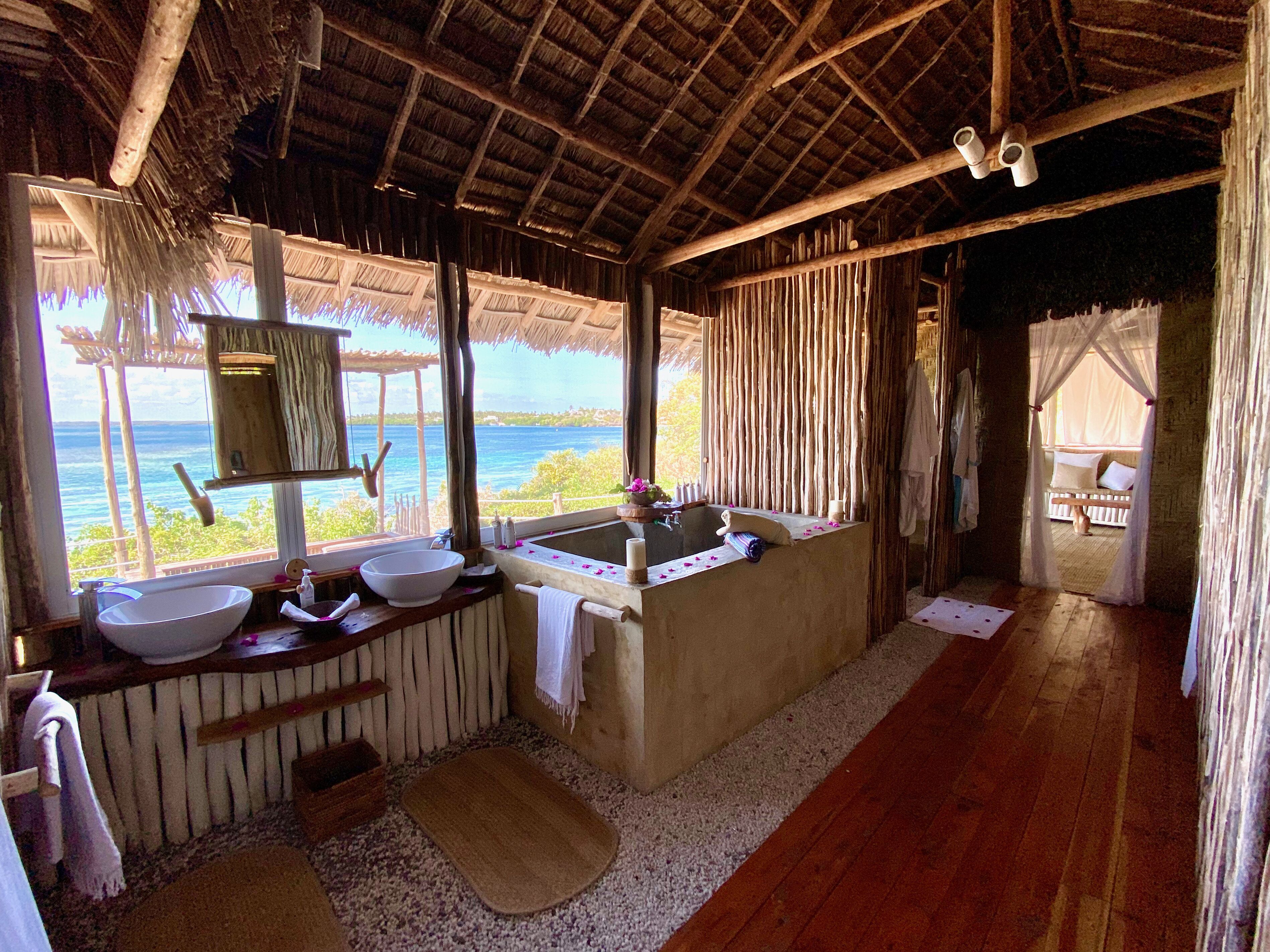 A bathroom with a large window overlooking the sea, a wooden floor, and a thatched roof.