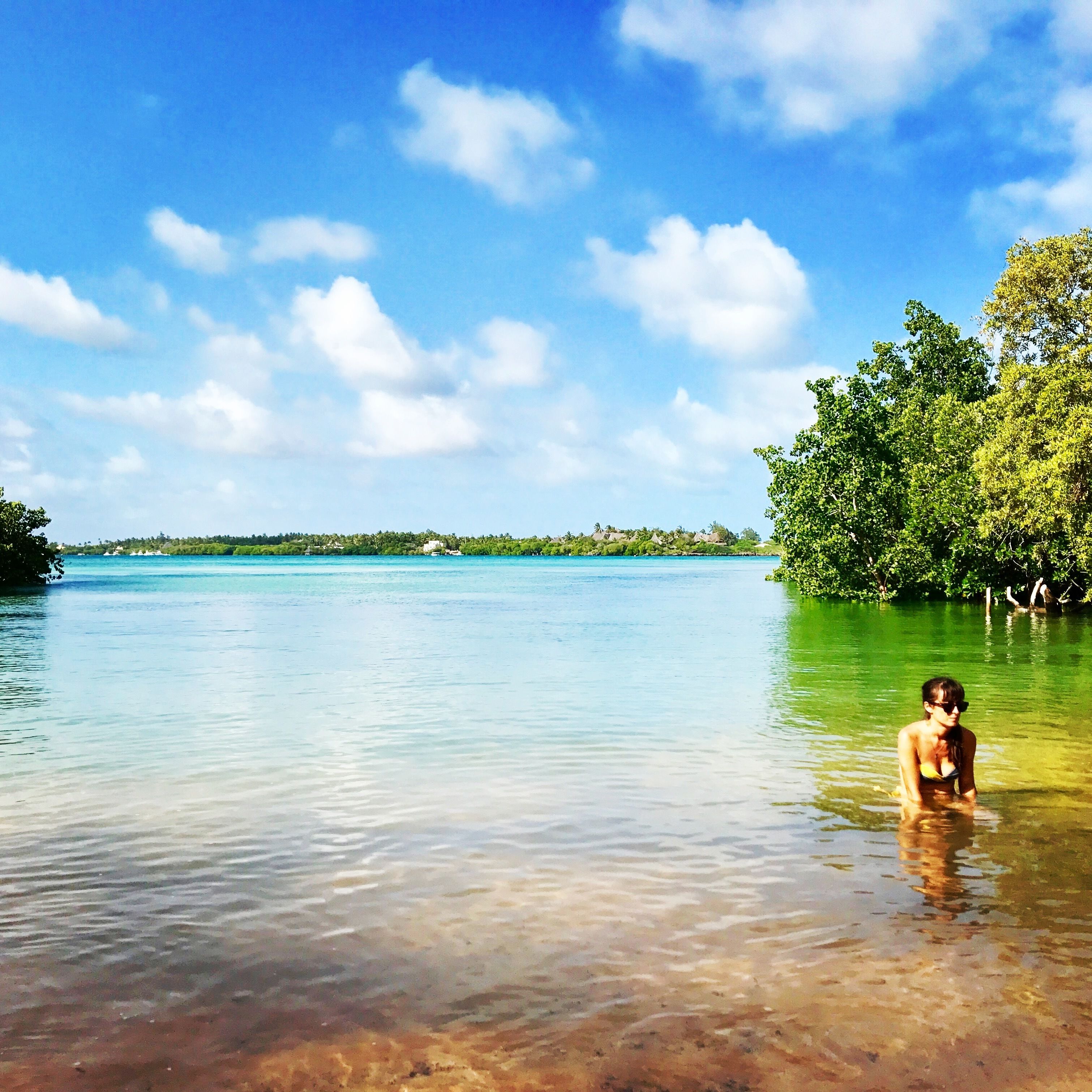 Eine Frau in einem Bikini im klaren, flachen Wasser mit Blick auf eine entfernte Küstenlinie und Bäume.