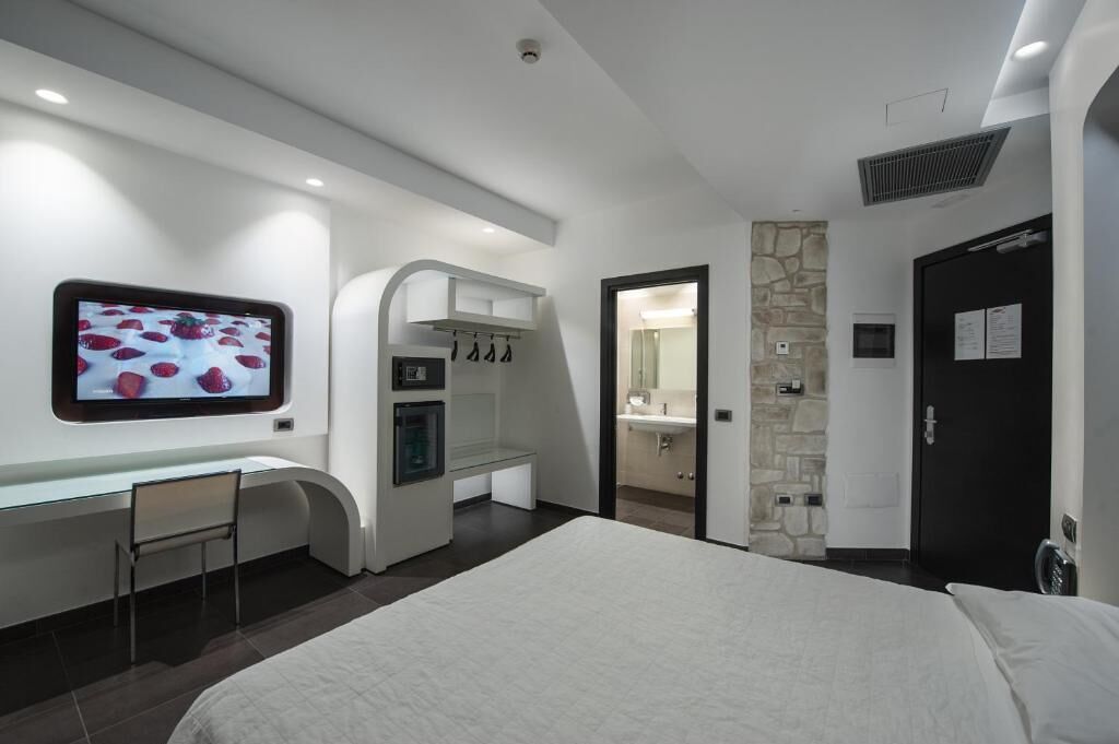 A modern hotel room with a bed, a desk with a chair, a TV mounted on the wall, a bathroom with a mirror, and a door.