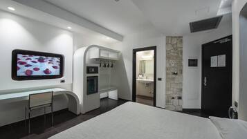 A modern hotel room with a bed, a desk with a chair, a TV mounted on the wall, a bathroom with a mirror, and a door.