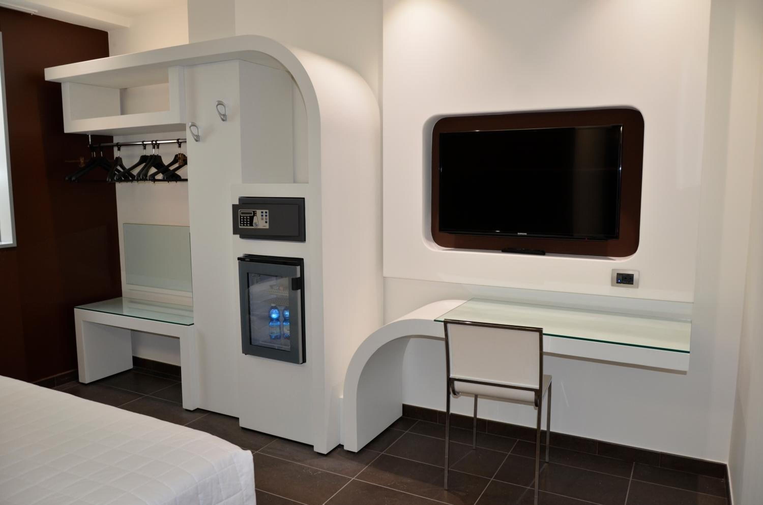 Una habitación de hotel moderna con una cama, un escritorio con superficie de vidrio, un televisor integrado, un minibar y un perchero.