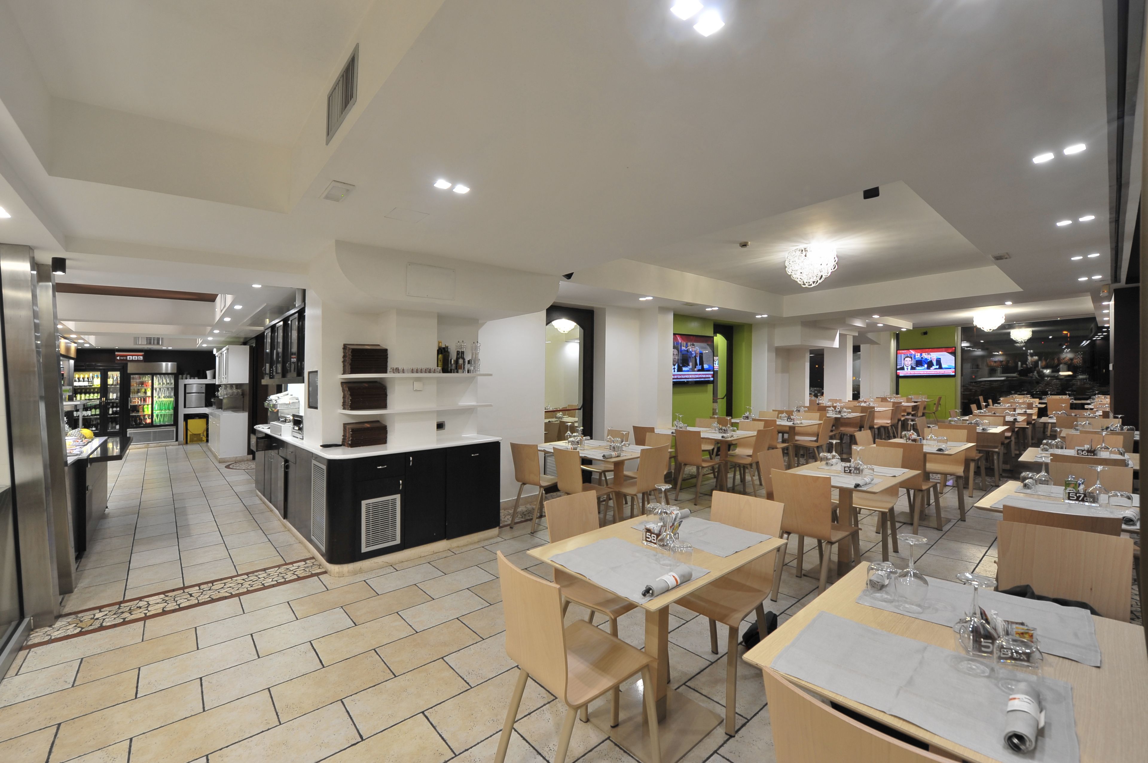 A restaurant with multiple dining tables and chairs, a kitchen area, and a television screen.