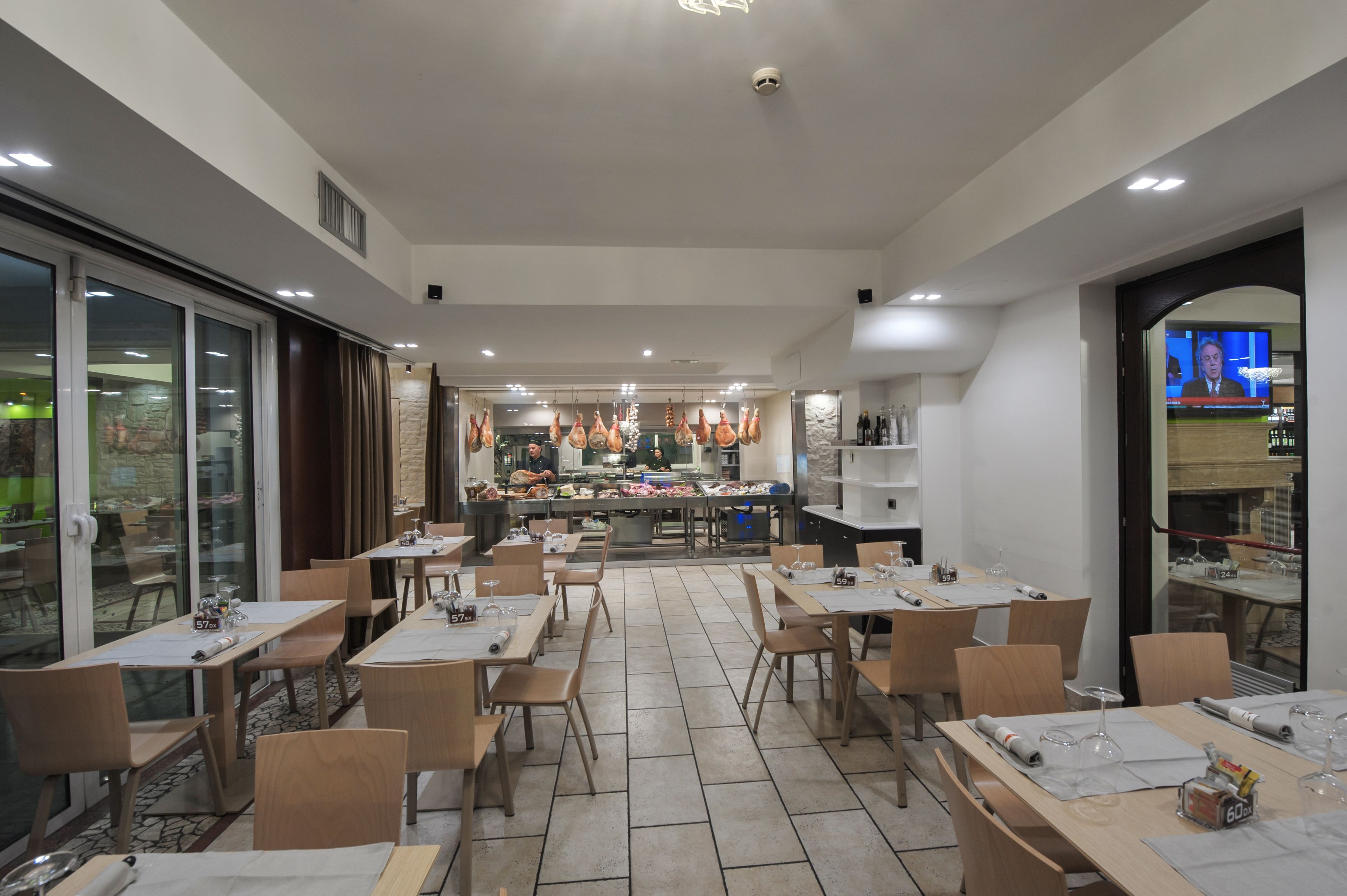 A restaurant with multiple tables and chairs, a kitchen area in the background, and a television screen displaying a news program.