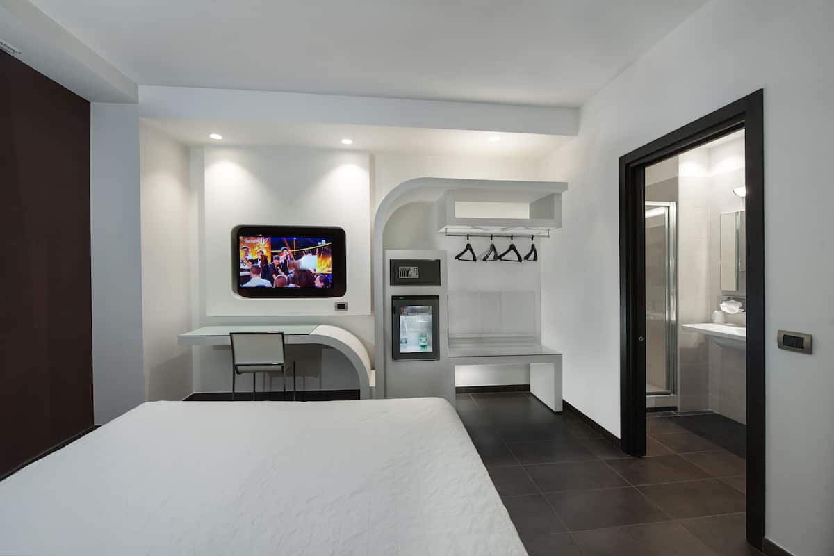 A modern hotel room with a bed, a desk with a chair, a TV mounted on the wall, a mini-fridge, and a bathroom visible through an open door.