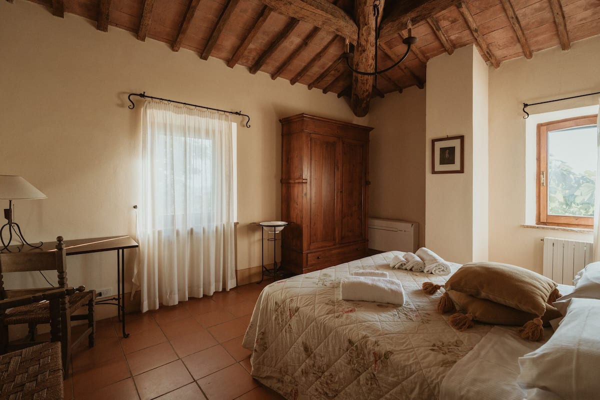 A bedroom with a wooden ceiling, a bed with a floral quilt, a wooden wardrobe, a desk with a lamp, and a window with curtains.