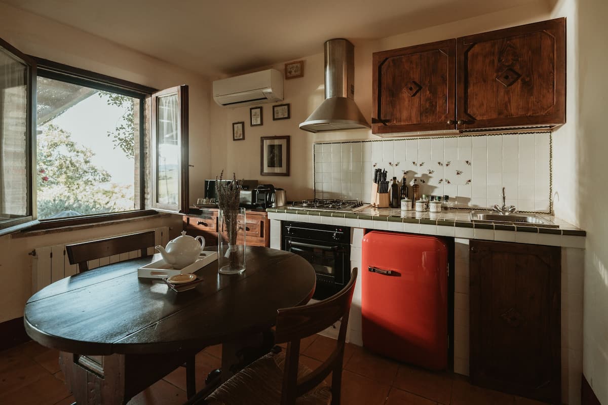 A kitchen with wooden cabinets, a red dishwasher, a window, and a table with a teapot.