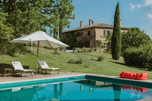 A pool area with lounge chairs, a white umbrella, and a house in the background. - Agriturismo Le Macchie (Pienza)