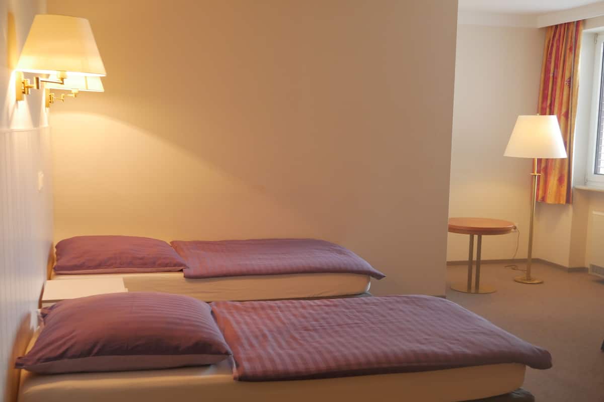 Une chambre d’hôtel avec deux lits, chacun agrémenté d’une literie violette, une petite table ronde et deux lampes fixées au mur.