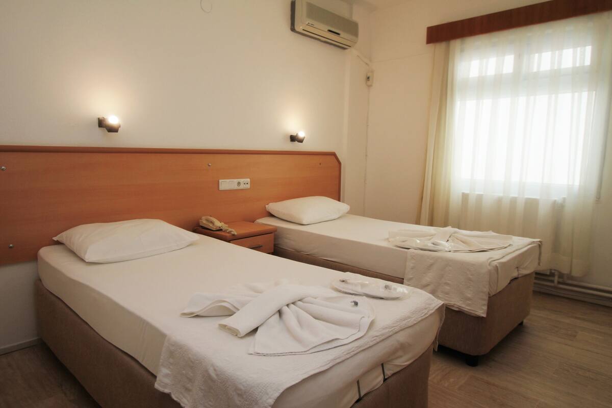 A hotel room with two beds, each with a pillow and a white blanket. There is a nightstand with a phone and a lamp. The room has a wooden headboard, a window with sheer curtains, and an air conditioning unit on the wall.
