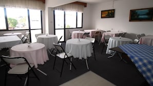 A dining area with round tables and chairs, large windows with curtains, and a painting on the wall.