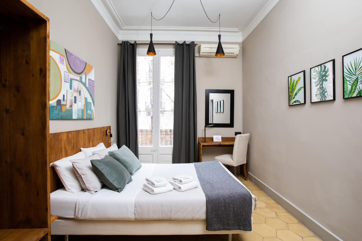 A hotel room with a bed, a desk, and a chair. There is a window with curtains, a painting on the wall, and a picture frame.