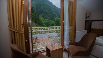 A room with a bed, two chairs, a small table, and a balcony with a view of a forested mountain.