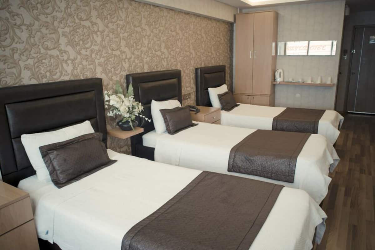 A hotel room with four beds, a nightstand with a vase of flowers, a cabinet, and a shelf with toiletries.