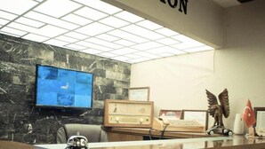 A reception area with a marble wall, a television mounted on the wall, a wooden reception desk, and a flag. - Yayla Hotel (Izmir)