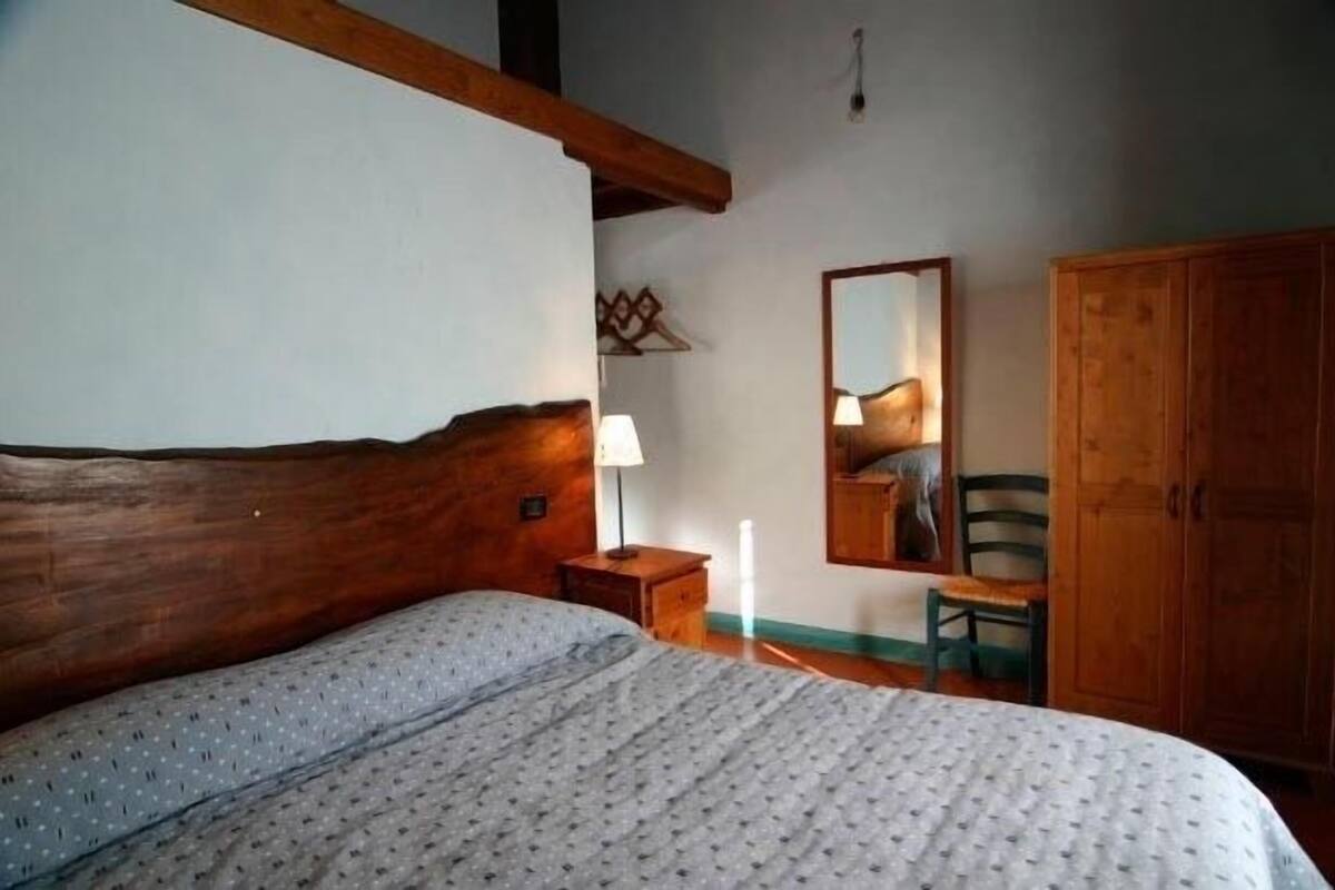 Een slaapkamer met een houten hoofdbord, een bed met een beddeken met een patroon, een nachtkastje met een lamp, een spiegel, een houten garderobe en een stoel.