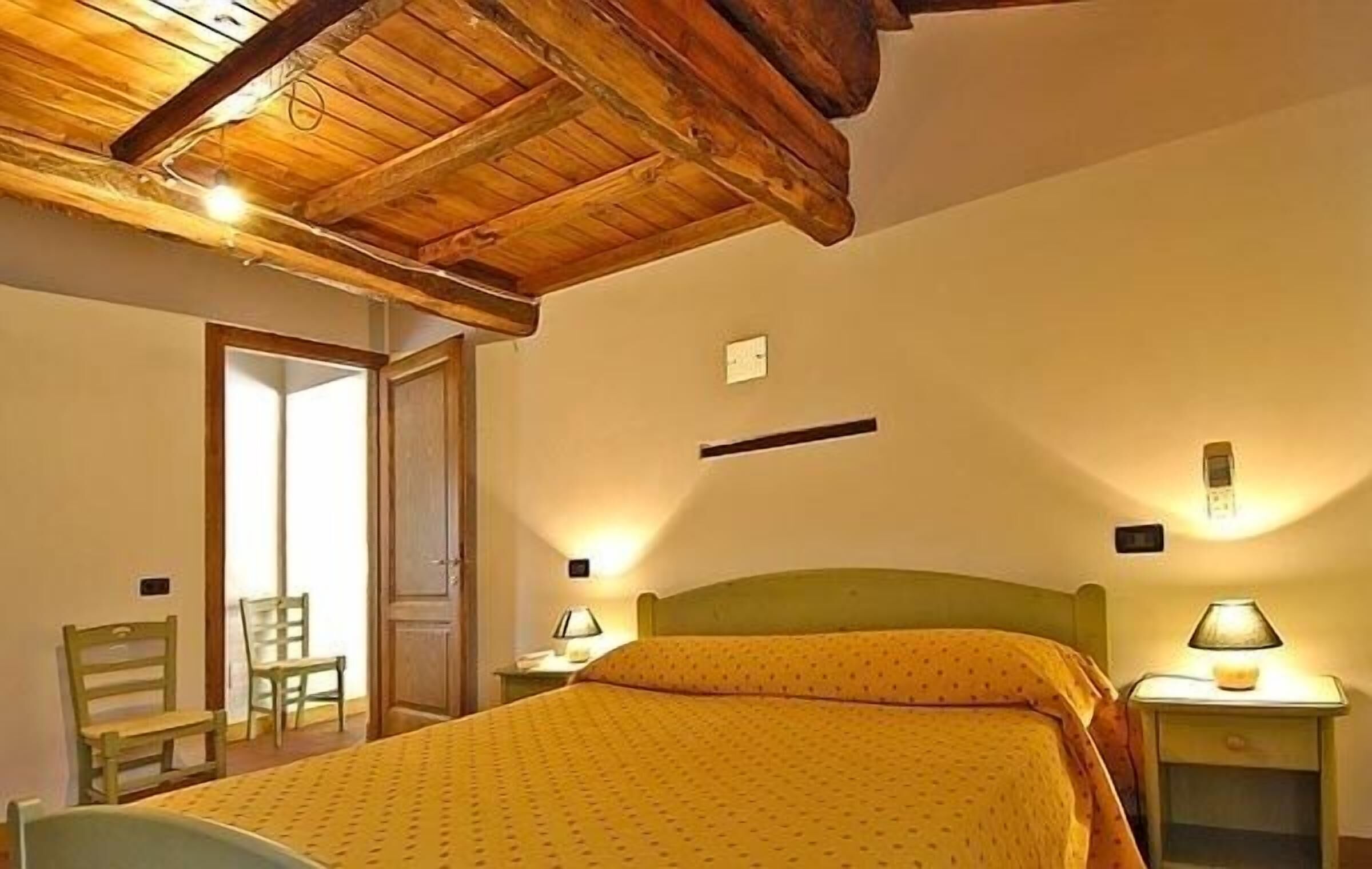 Une chambre avec un plafond en bois, un lit recouvert d’une couverture jaune, deux tables de chevet avec des lampes et une chaise.
