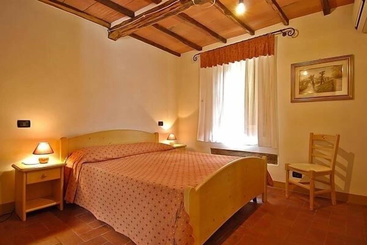 Een slaapkamer met een houten bed, twee bedlampen, een stoel, een raam met gordijnen en een ingelijste foto aan de muur.