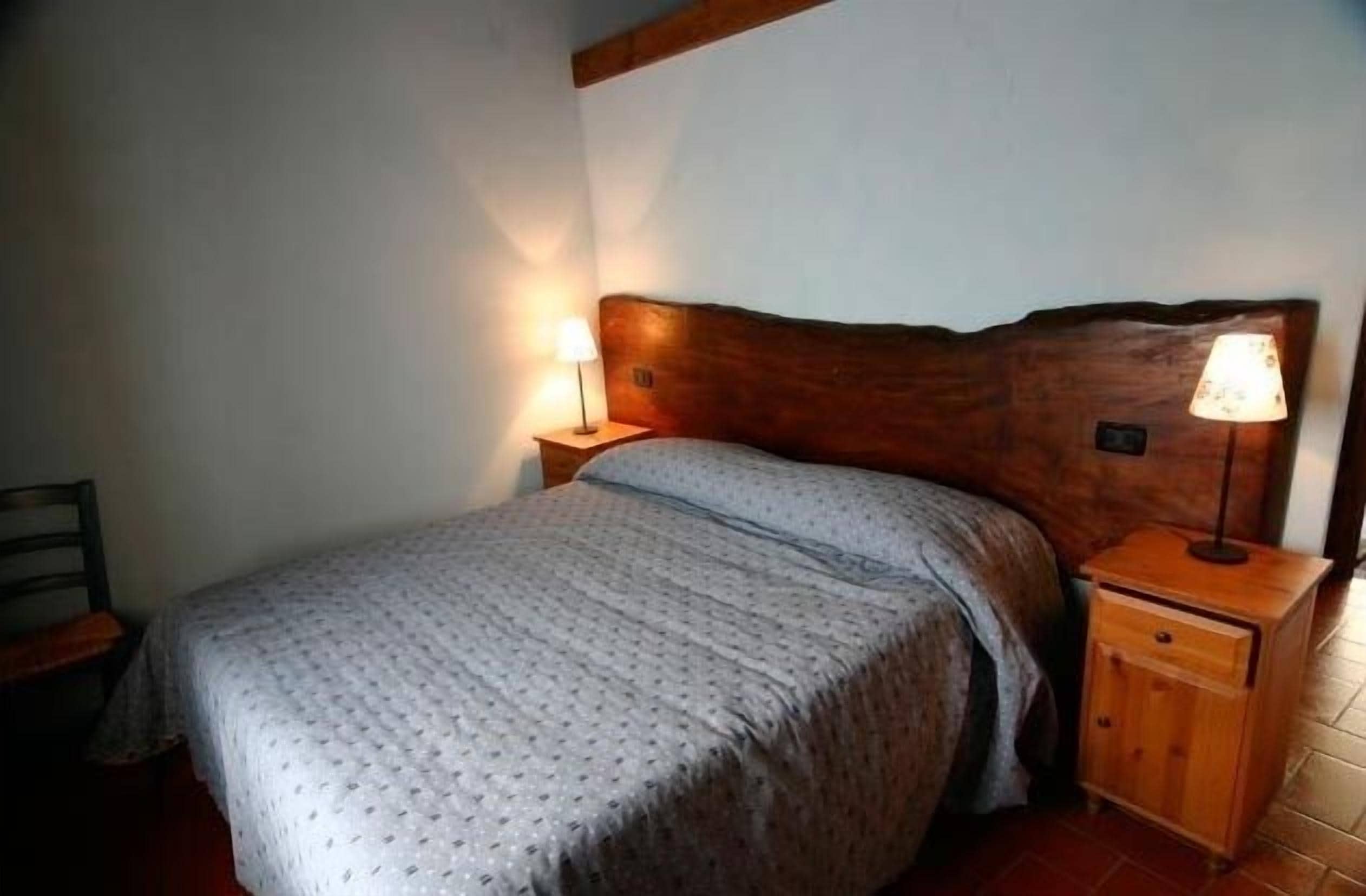 A bedroom with a wooden headboard, a single bed with a patterned bedspread, two bedside lamps, and a nightstand with a drawer.
