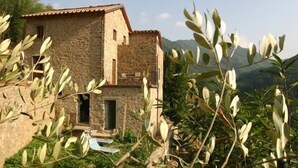 Un bâtiment en pierre, doté d’un toit en tuiles, entouré de verdure et avec des montagnes en arrière-plan.