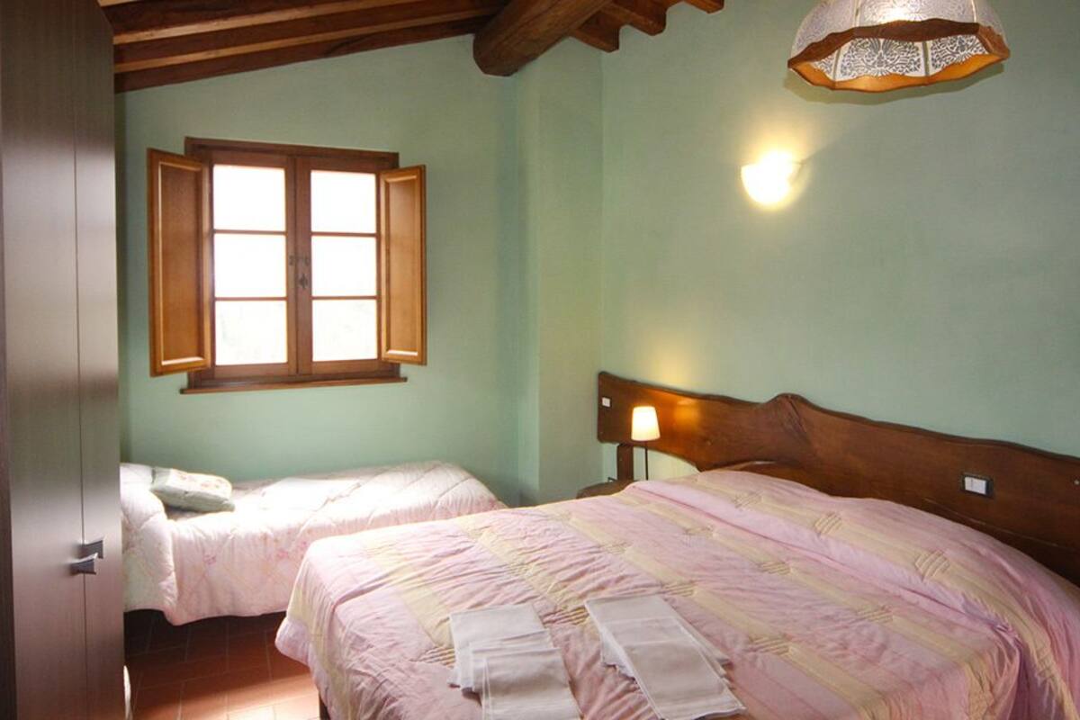 Een slaapkamer met twee bedden, houten hoofdeinden, een raam met open luiken en een wandlamp.