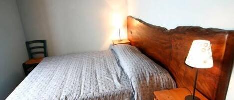 Een klein, net opgemaakt bed met een houten hoofdbord en een nachtkastje met een lamp.