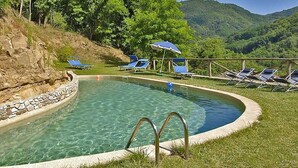 Une piscine aux eaux cristallines, bordée de pierres, entourée de chaises longues et de parasols, le tout sur fond de paysage montagneux.