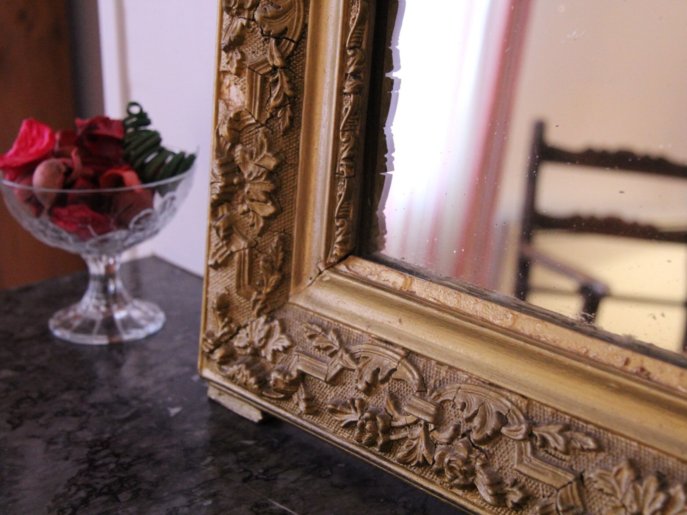A gold-framed mirror with floral carvings, a glass bowl with red roses, and a dark marble surface.