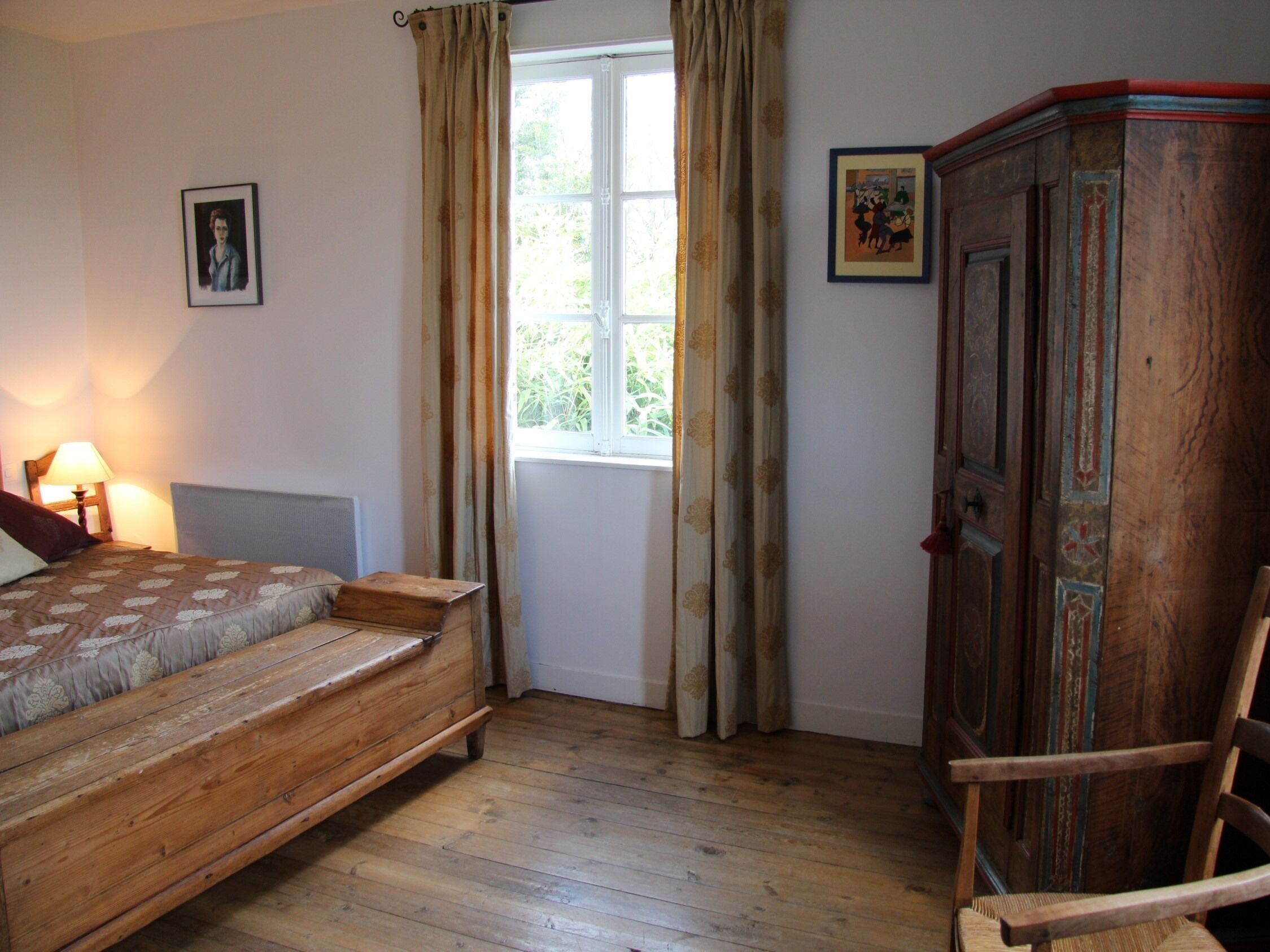 Un dormitorio con una cama de madera, una mesita de noche, una lámpara, un ventanal con cortinas y un armario de madera.
