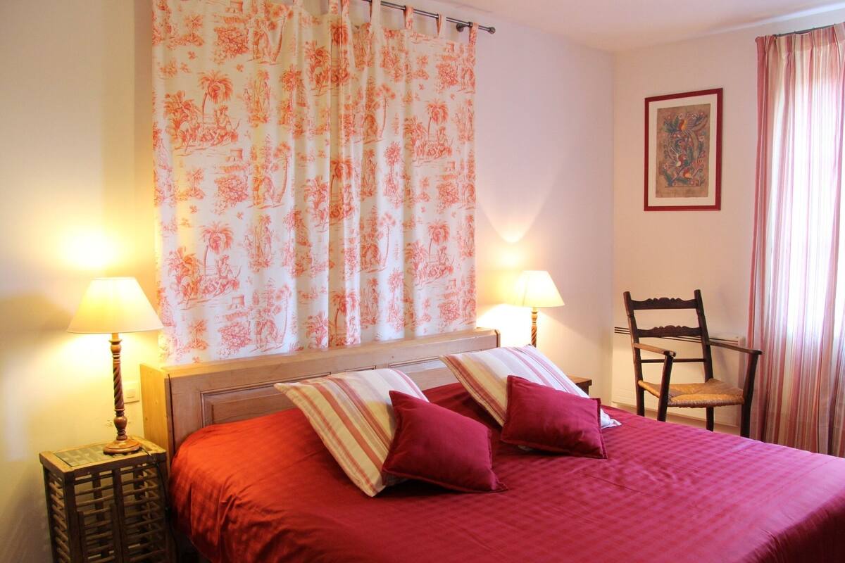Un dormitorio con cama, dos lámparas de noche, una silla, una mesita de madera y una cortina con estampado floral.