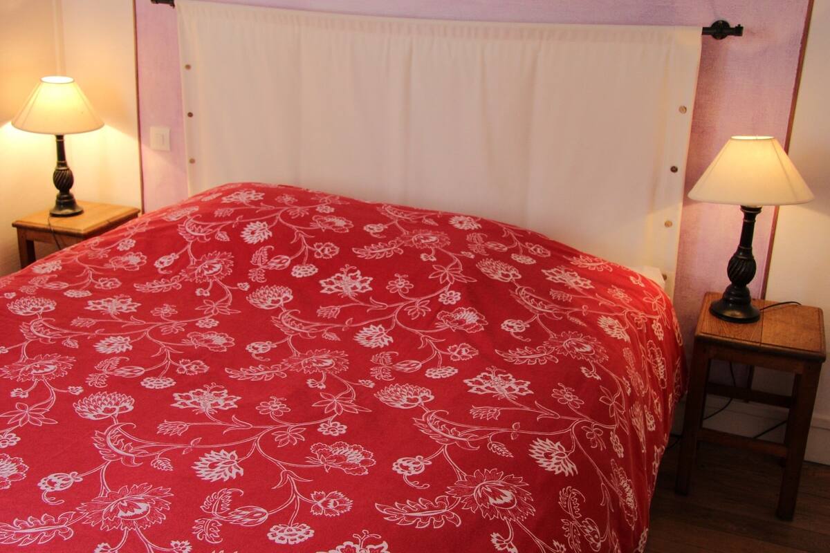 Una cama con un edredón rojo estampado con motivos florales, dos lámparas de noche y un cabecero blanco.