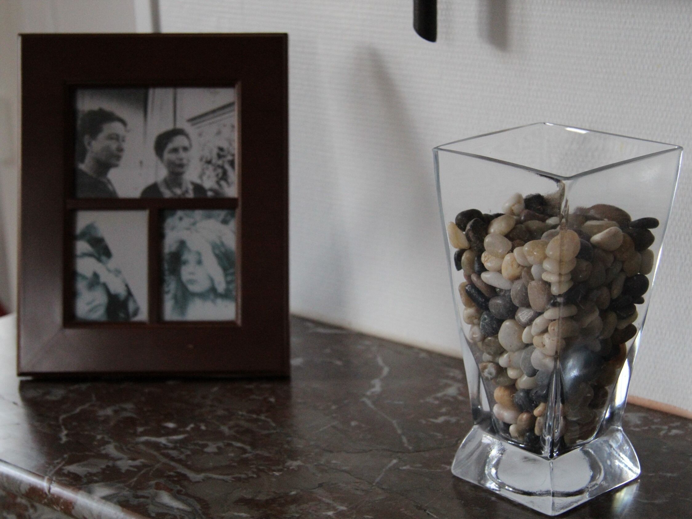 A glass vase with pebbles on a marble countertop, next to a framed photograph.