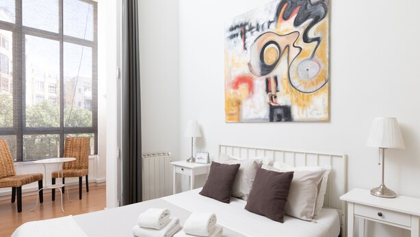 A hotel room with a large bed, two pillows, a bedside table with a lamp, a chair, and a window with a view of buildings. - CasaNova Guest House (Barcelona)