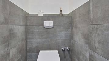 Et moderne badeværelse med et hvidt toilet, grå fliser og en hylde med toiletartikler.