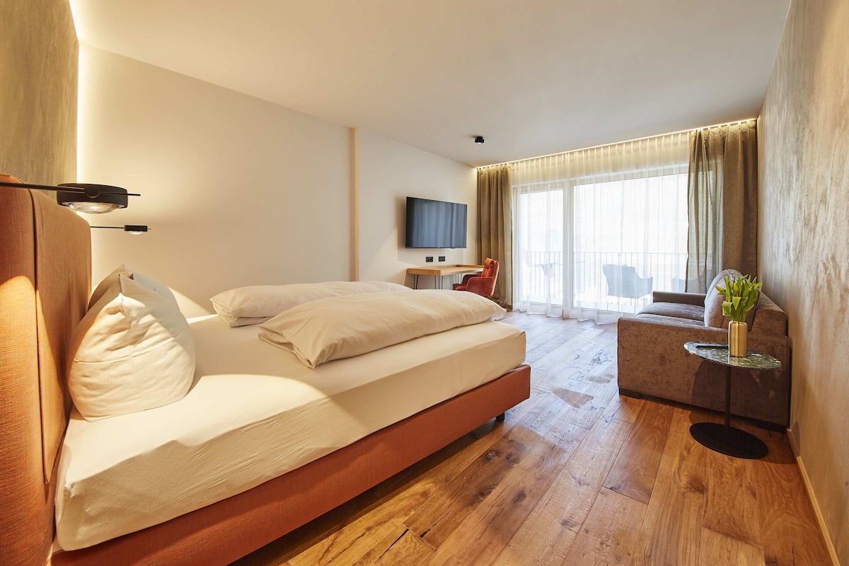 Quarto de hotel moderno com cama grande, pequena escrivaninha, televisão e varanda com sofá e cortinas.
