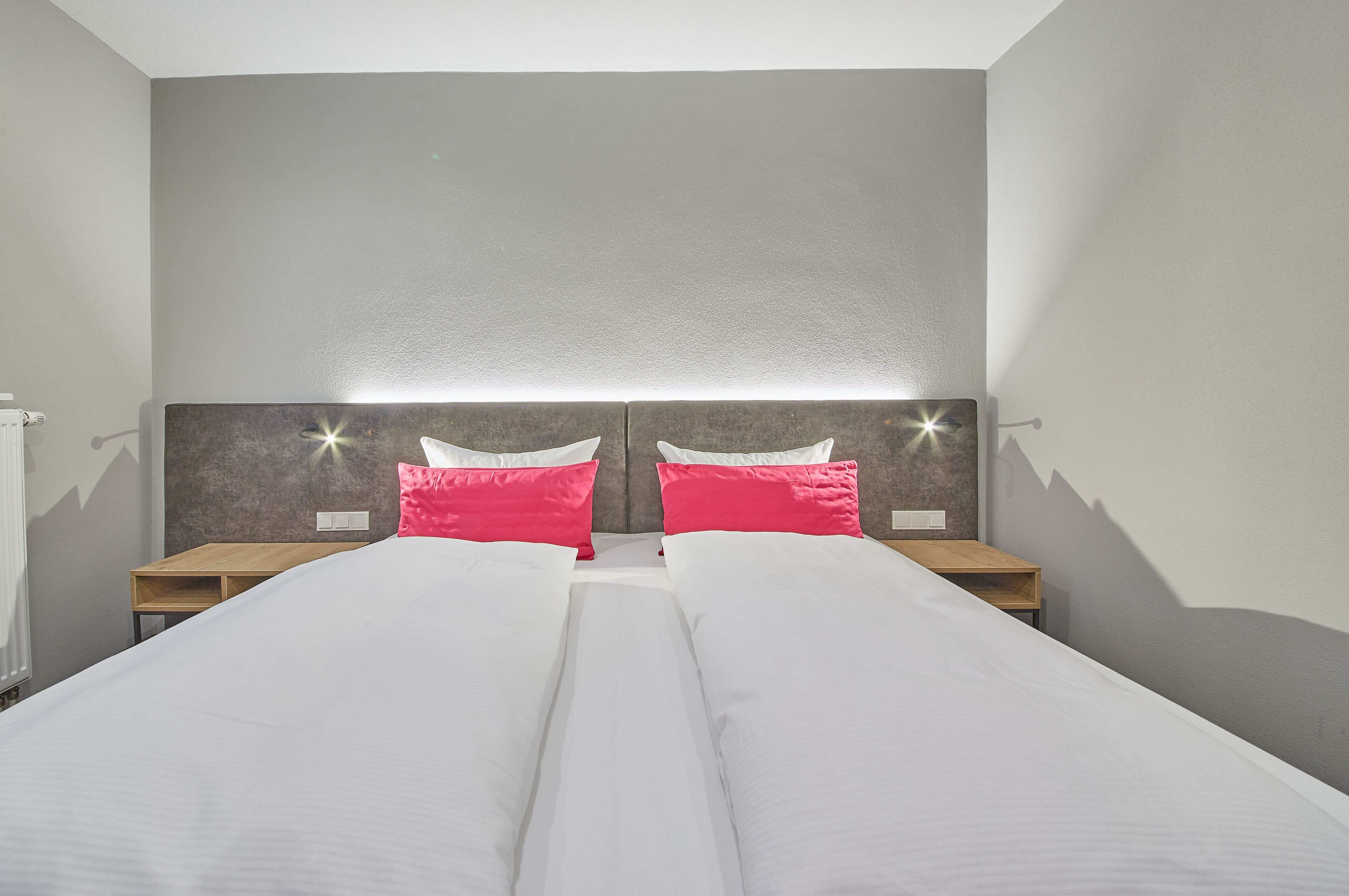 Un lit double avec une literie blanche et rose, deux tables de chevet et un mur gris à texture.