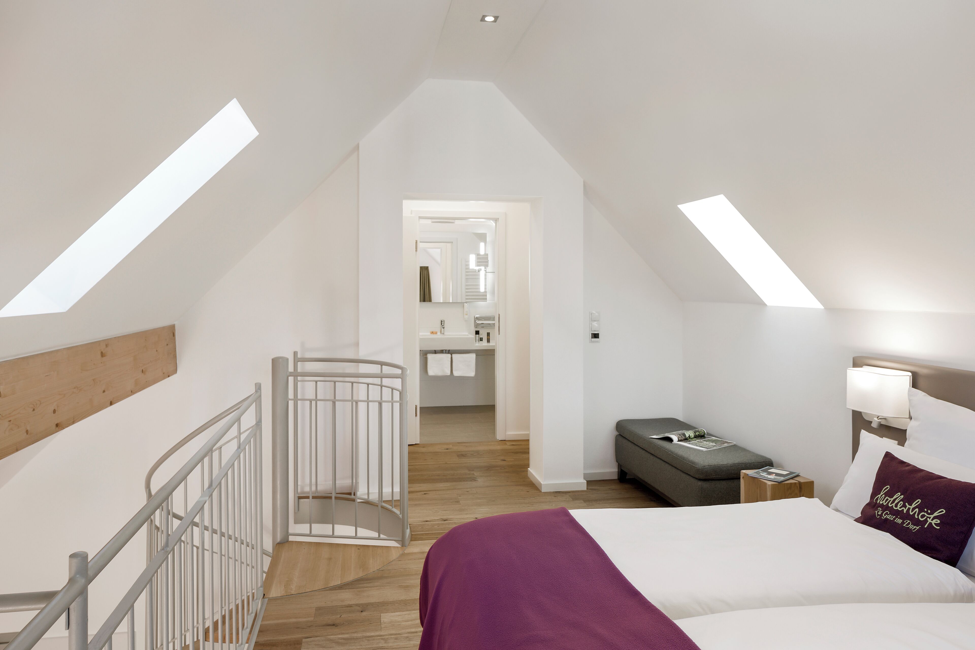 Une chambre avec un plafond en pente, des fenêtres de toit, un lit recouvert d’un plaid violet, une table de chevet avec une lampe et un petit coin salon.