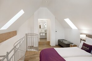 A bedroom with a sloped ceiling, skylights, a bed with a purple throw, a nightstand with a lamp, and a small seating area.