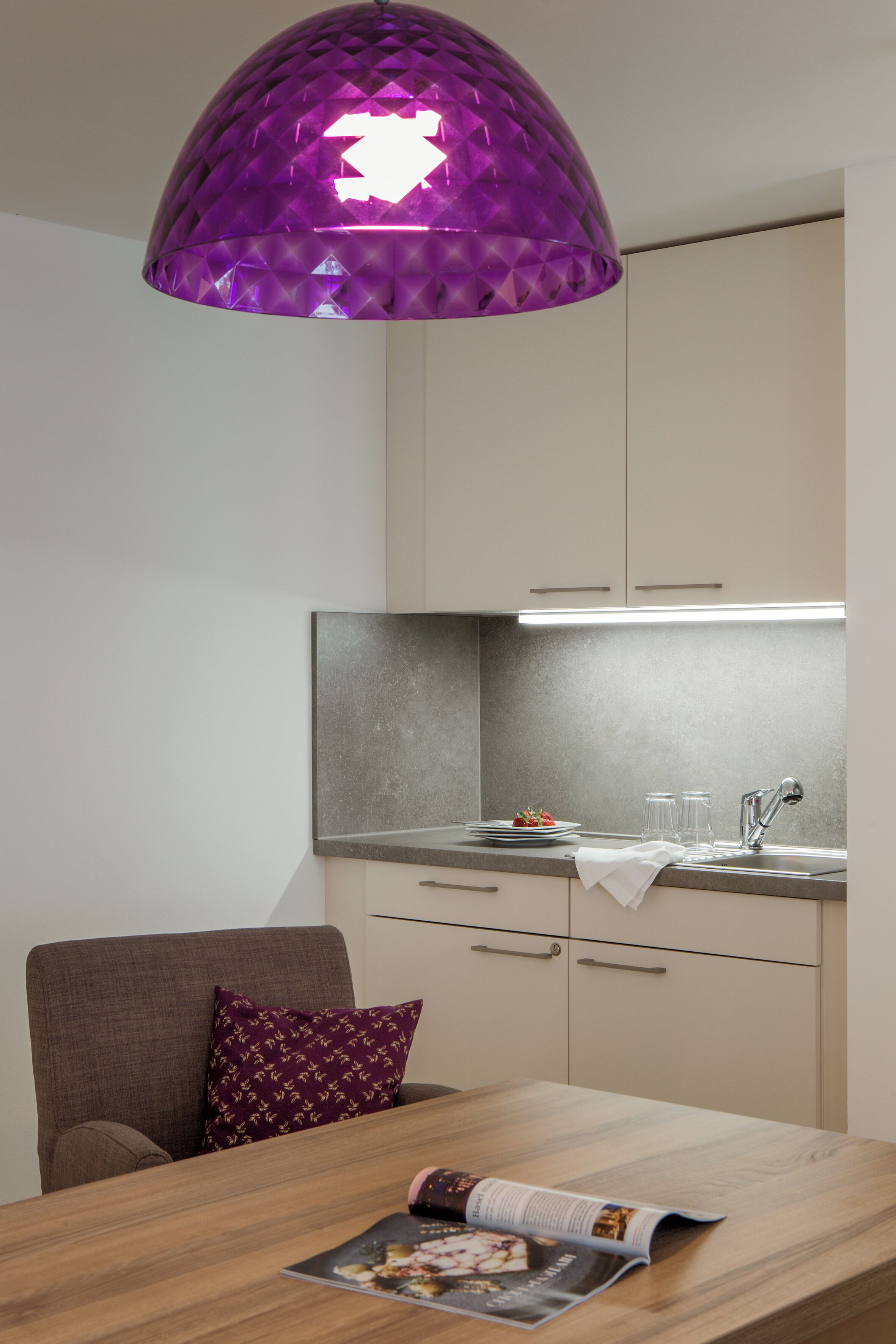 Une cuisine moderne avec un luminaire suspendu violet, une table à manger en bois et un magazine.