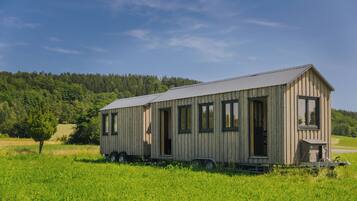 Une cabane en bois dotée d’une grande fenêtre et d’une porte, située dans un champ herbeux, avec une forêt en arrière-plan.