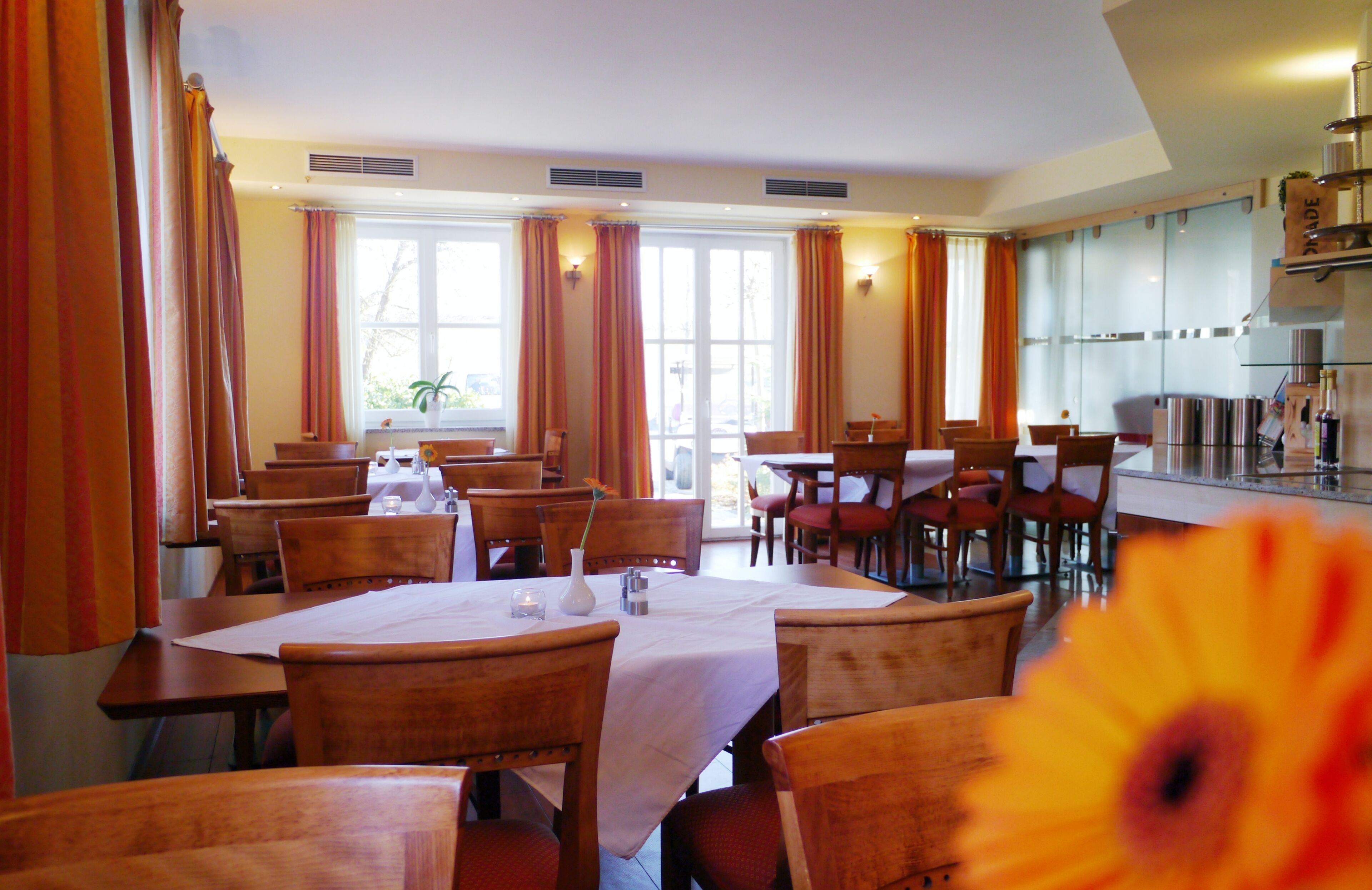 Zona pranzo con diversi tavoli e sedie, ampie finestre con tende e angolo cottura sullo sfondo.