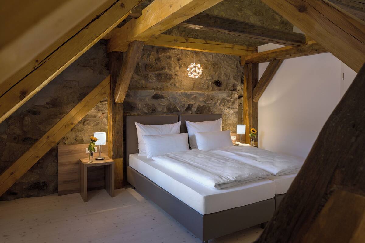 Une chambre avec un mur en pierre, des poutres apparentes, un lit avec une literie blanche et deux tables de chevet avec des lampes.