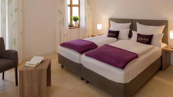 A double bed with white and purple bedding, two bedside tables with lamps, a window with a plant, and a wooden side table with a book.