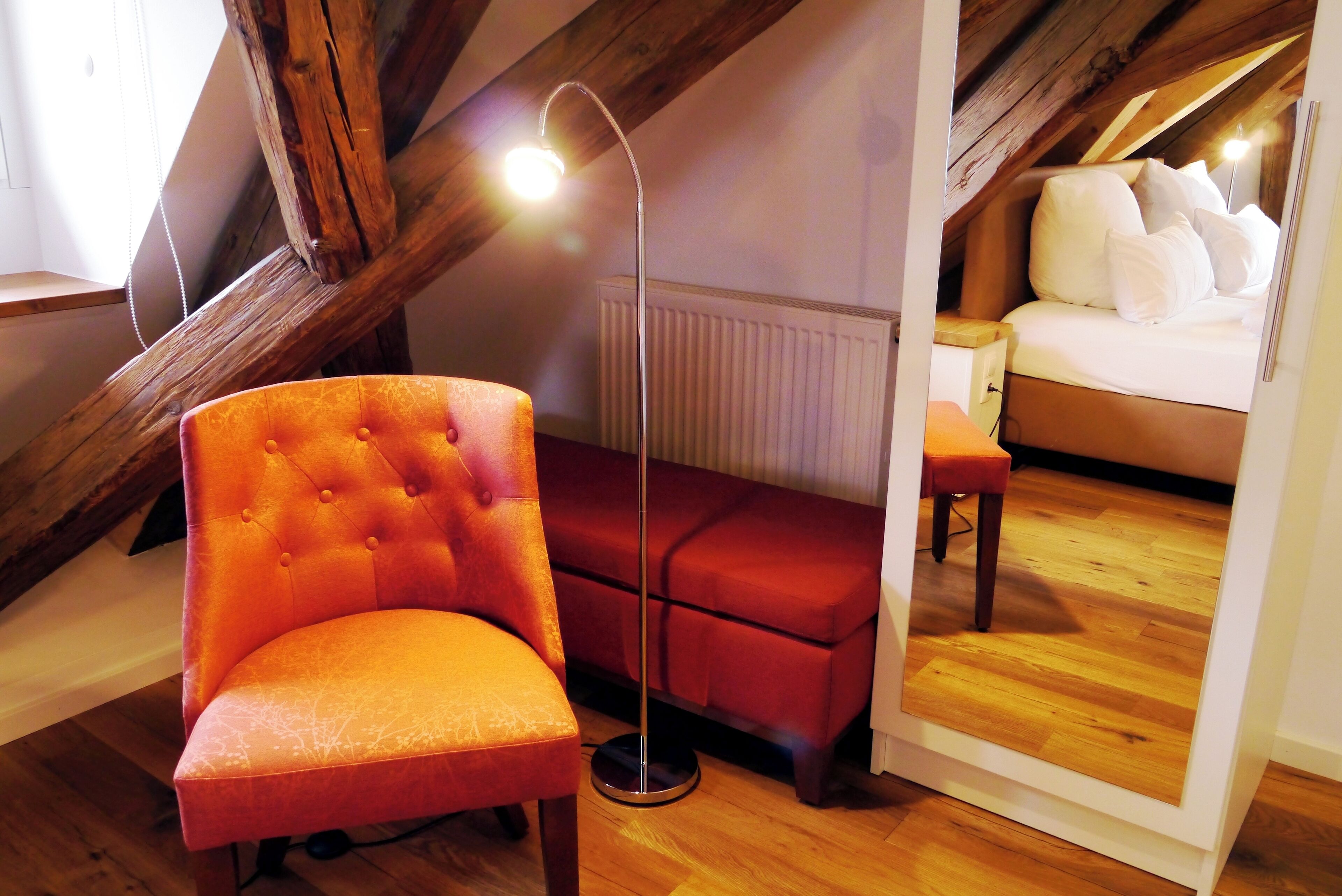 Un dormitorio acogedor en el ático, con techo de madera, un sofá rojo, un sillón naranja con capitoné, una mesita de noche y una cama con ropa blanca.