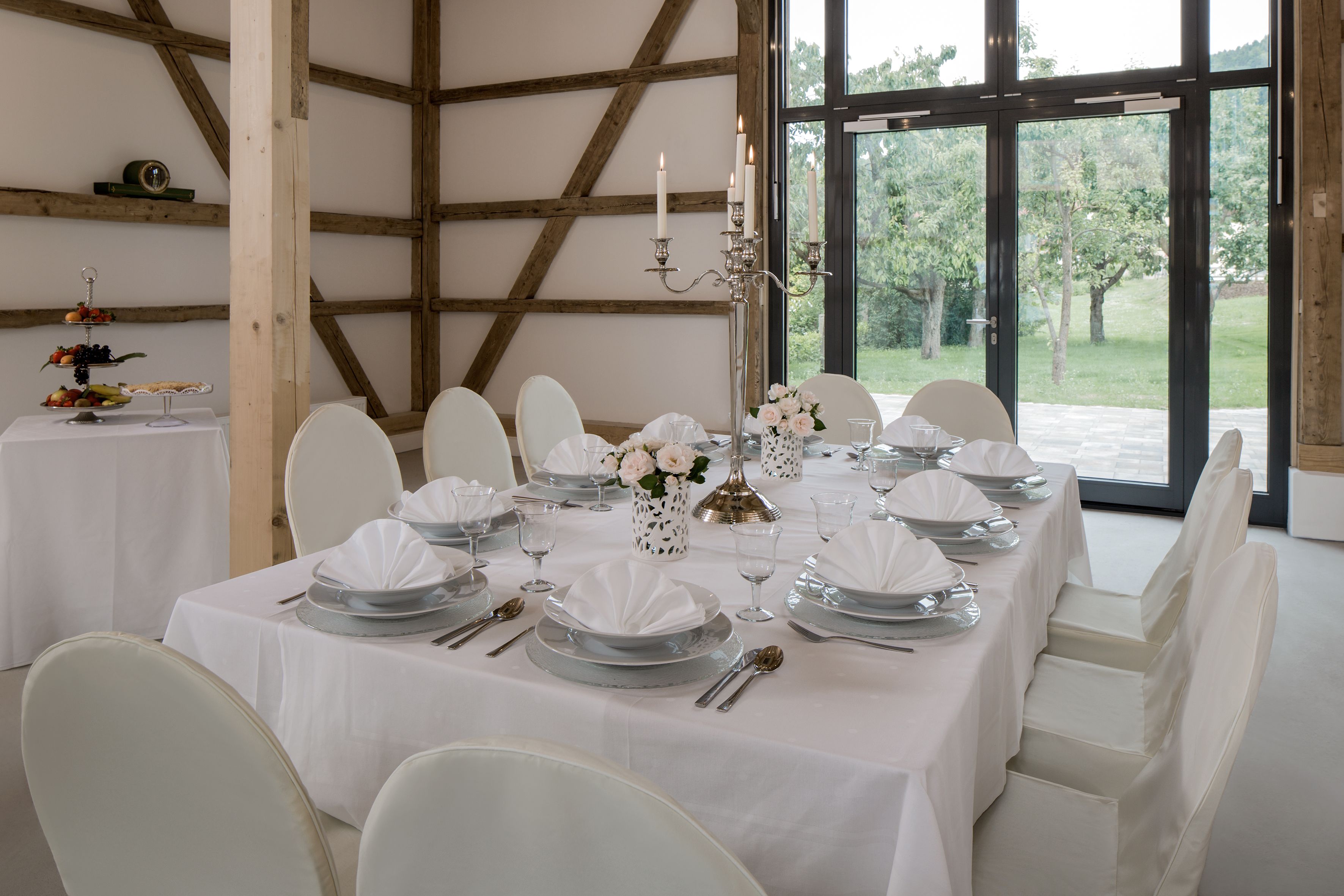 Uma sala de jantar com uma mesa comprida posta para refeição, cadeiras brancas e vista para o verde através de grandes janelas.