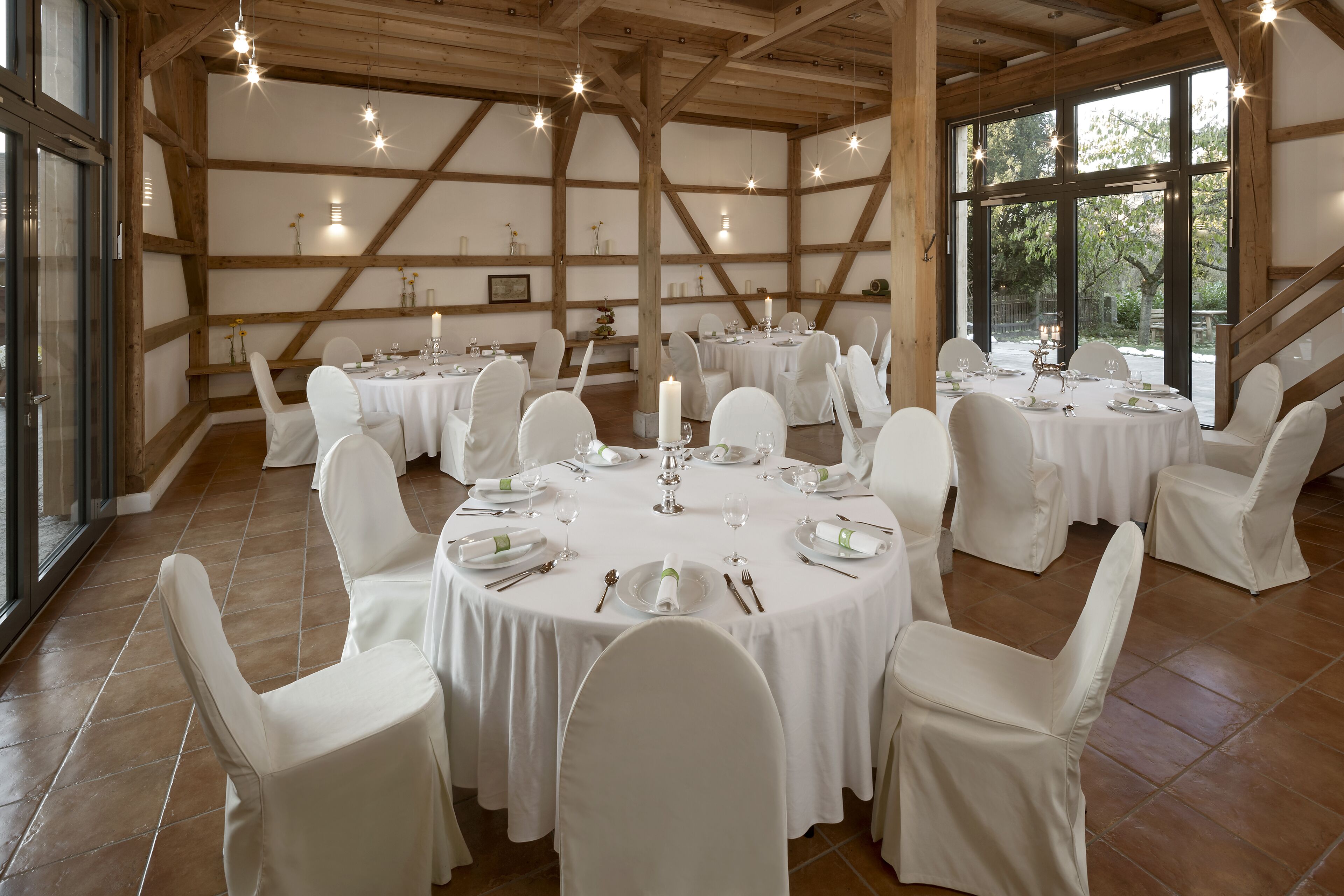 A large hall with multiple round tables set for dining, white chairs, and a rustic wooden ceiling.