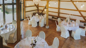 A spacious hall with high wooden beams, round tables set for dining, and a chandelier.