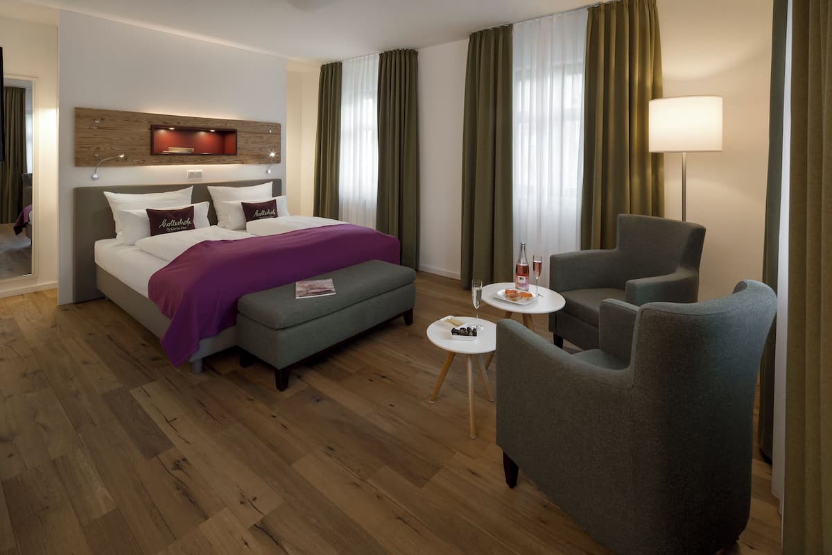 Une chambre d’hôtel avec un lit, un fauteuil gris, une petite table ronde avec une bouteille et un verre, et une lampe.