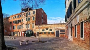 A courtyard with stone benches, a small tree, and buildings with brick facades and multiple windows. - Domus Orsoni (Venice)
