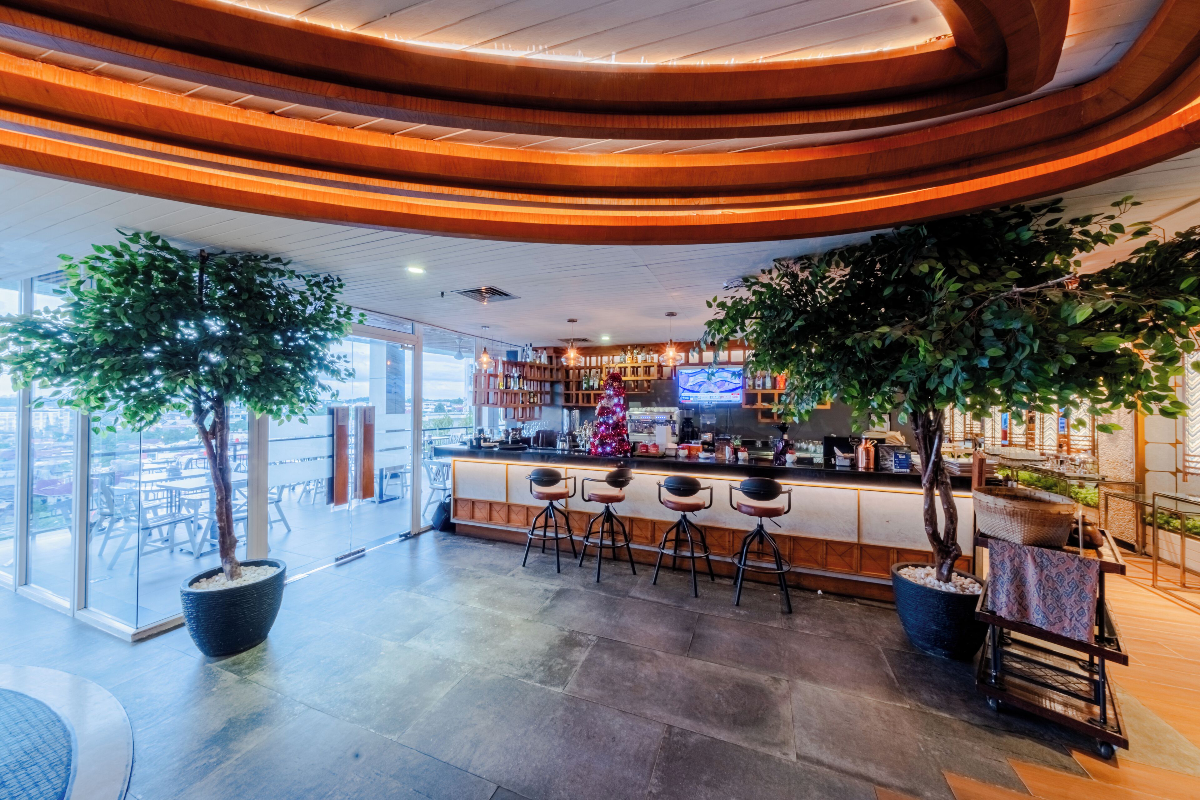 A modern bar area with a curved wooden ceiling, a large potted tree, and a television screen.