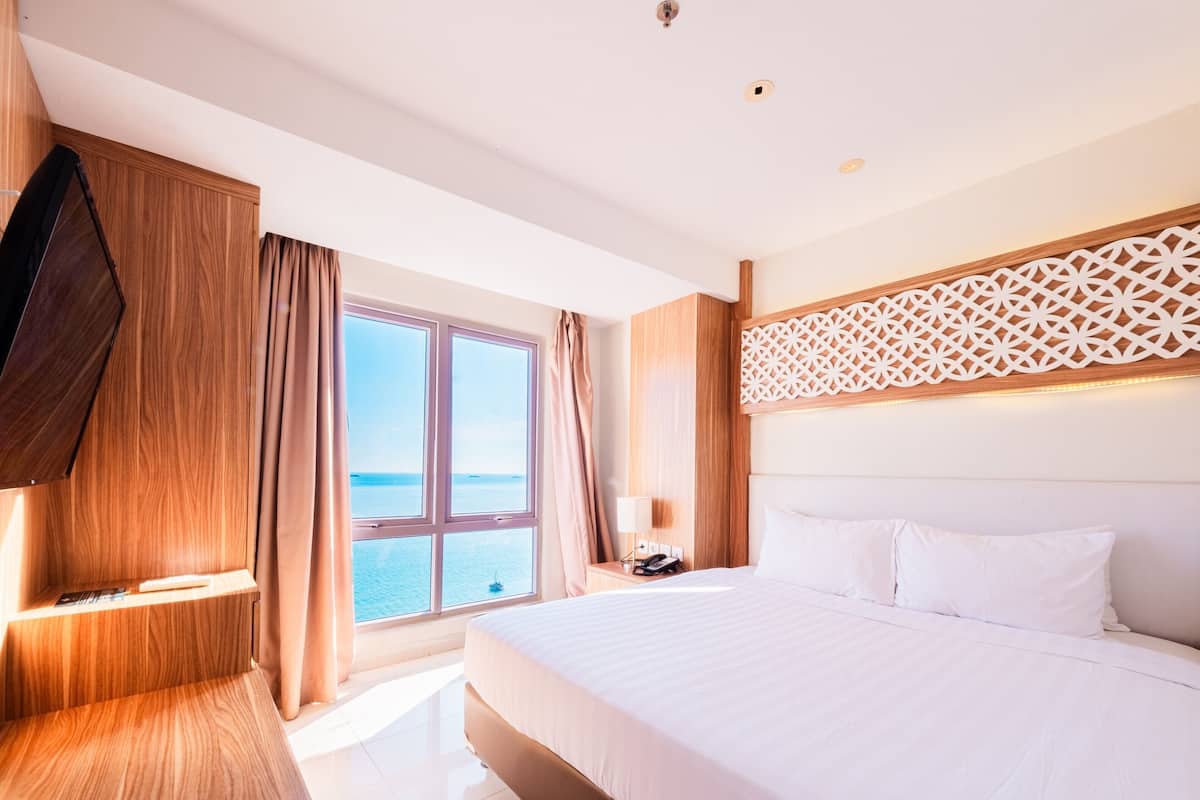 Een hotelkamer met een groot bed, een aan de muur bevestigde televisie, een nachtkastje en uitzicht op zee door een groot raam.